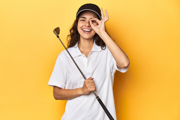 Golfer woman with cap, golf polo, yellow studio, excited keeping ok gesture on eye.
