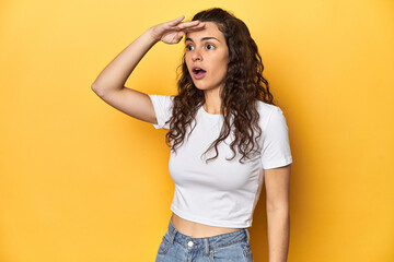 Young Caucasian woman, yellow studio background, looking far away keeping hand on forehead.