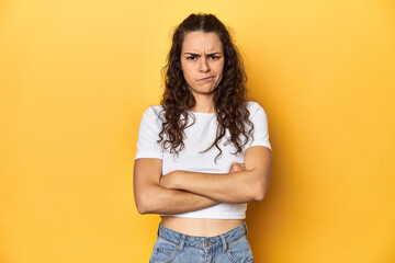 Young Caucasian woman, yellow studio background, frowning face in displeasure, keeps arms folded.