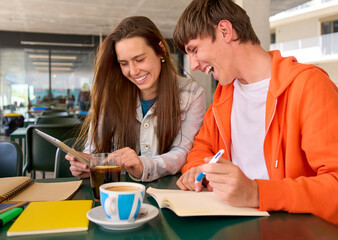 Obraz premium Young Caucasian female and male laughing while using tablet and notepad in university campus cafeteria. College classmates taking notes and drinks at break time. Concept of generation z student people