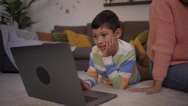 Smiling Latin Little Boy Watching Funny Videos On Laptop Lying On Living Room Carpet. Happy Mother And Son Hanging Out Using Technology At Home. Family Relationships And Parental Care In Childhood.