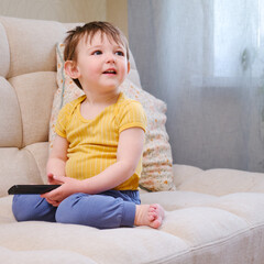 Fototapeta premium Toddler watches their favorite cartoon on TV while sitting comfortably on the sofa. The smiling child watches cartoons on TV, sitting on the beige sofa. Kid aged about two years (one year ten months)