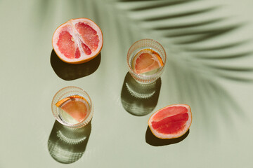 Summer flat lay with two glasses of refreshment drinks and grapefruit slices on green background with palm leaf shadow sun and sunlight. Vacation, holiday, mocktail, creative minimal concept.