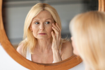 Skin aging concept. Beautiful aged woman looking at mirror at wrinkles near eyes, checking face,...
