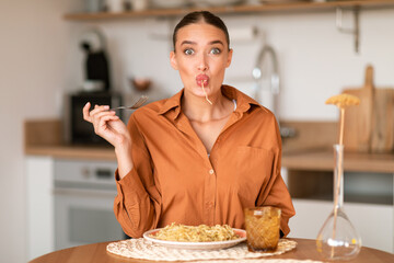 Joyful culinary moments. Funny emotional lady tasting delicious homemade spaghetti, sitting at table in kitchen interior