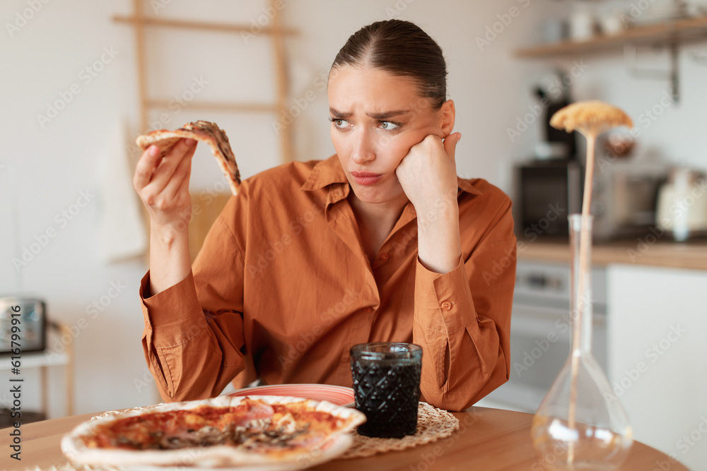 Wall mural upset woman looking at delicious piece of high-calorie pizza, holding slice in hand