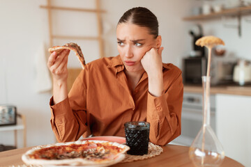Upset woman looking at delicious piece of high-calorie pizza, holding slice in hand