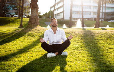 Calm young businessman in formal wear meditating with closed eyes outdoors, practicing yoga, sitting on lawn in park