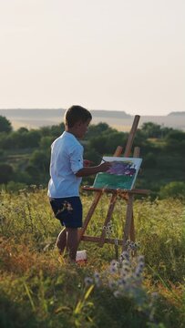 Vertical Screen: Little boy wearing white shirt standing in front of wooden easel and painting on canvas at sunset on hill. Creative hobby. Concept of arts