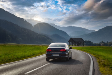 Travel. Car rides on an asphalted mountain rural road in a Slovenian village. Beautiful landscape with passenger machine, curved highway, alpine mountains, hills, fields green grass at dawn in summe