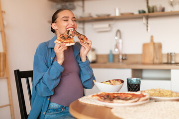 Excited young pregnant lady enjoying pizza, biting two tasty slices, sitting at table in kitchen...