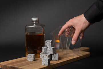 Squared whisky bottle on a wooden serving trey with icelandic granite whisky stones and and diamond prism whisky glasses being picked up by bartender. Black background.