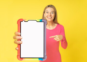 young friendly attractive woman with a joyful face holds mock-up screen tablet with cover in her hands and finger pointing on white isolated yellow background. Emotionaly happy face and promo notes.