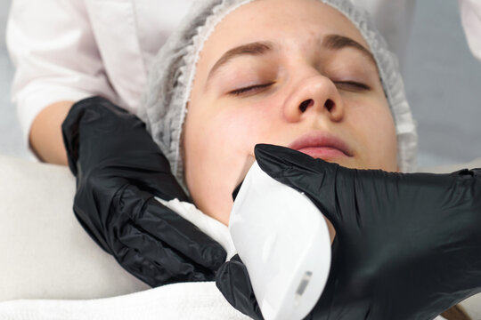 Cosmetologist making facial treatment with ultrasonic spatula to young woman. Ultrasound facial peeling at cosmetology clinic. Health care therapy with selective focus