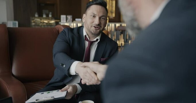 Over the shoulder shot of two businessmen reading document clanging coffee cups and shaking hands in hotel lobby. People and cooperation concept.