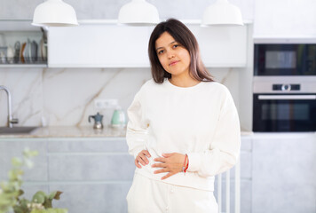 Fototapeta na wymiar Smiling young brunette in white casual clothes standing in cozy light home kitchen interior..