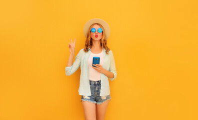 Portrait of young woman posing blowing her lips with smartphone wearing summer straw hat and shorts on yellow background