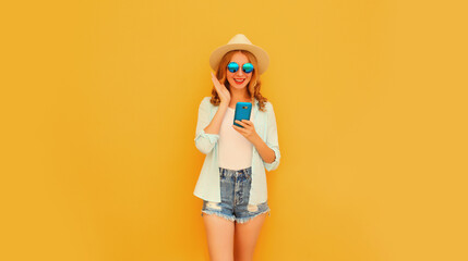 Happy smiling young woman 20s with smartphone wearing summer straw hat, shorts on yellow background