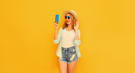 Portrait of happy smiling young woman taking selfie with smartphone or talking on video call waving hand wearing summer straw hat, shorts on yellow background