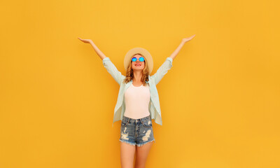 Happy cheerful caucasian smiling young woman raising her hands up wearing shorts, summer straw hat on yellow background