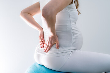 Pregnant woman with baby bump feels back pain and holds hands on her aching back while sitting on...