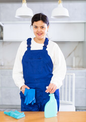 Portrait of young smiling cleaning lady in blue overalls standing near table in light interior of home or office kitchen, holding rag and detergent spray. Woman worker ready to clean up
