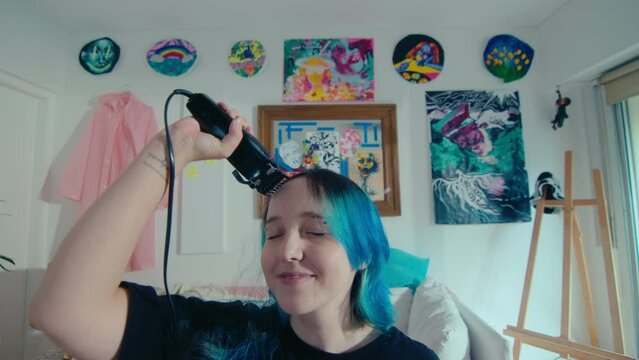 Young girl with blue hair shaving her head with electric razor, looking at camera, smiling and enjoying new hairstyle. Medium close-up shot, video portrait