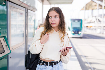 Confident girl with a backpack on her back, walking along a tram stop, holds a mobile phone in her...