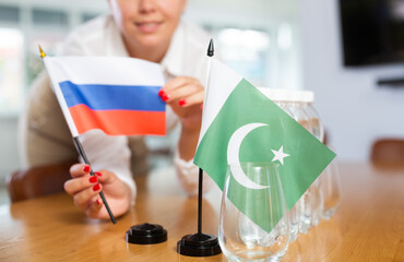 Positive young woman putting little flag of Russia on table next to the flag of Pakistan and...