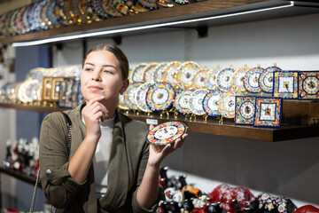 Young woman tourist in casual clothes chooses decorative clock in souvenir shop