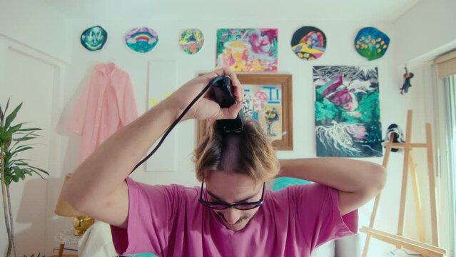 Young handsome guy with mustache and beard wearing trendy narrow sunglasses and pink t-shirt shaving head with electric razor when doing self-haircut at home