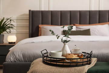 Wicker tray with vase of eucalyptus leaves, tea and cookies on ottoman in bedroom. Interior design