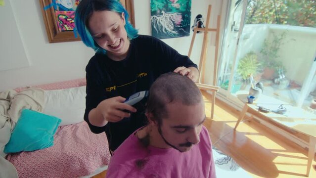Young happy girl with blue hair shaving hair of her boyfriend with electric razor, smiling when giving haircut at home. High angle view