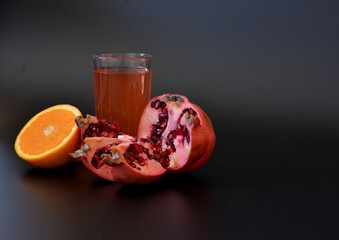 Tall glass with orange juice and pomegranate on a black background, next to the halves of ripe fruit.