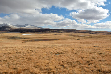 Fototapeta na wymiar A huge flat steppe with dry, yellowed grass at the foot of snow-covered mountain ranges on a sunny autumn day.