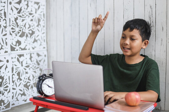Focused Asian schoolboy use laptop for online learning class and raises his hand to answer lesson. Home schooling and online virtual education concept
