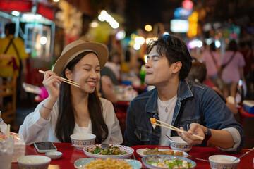 Young Asian couple traveler tourists eating Thai street food together in China town night market in...