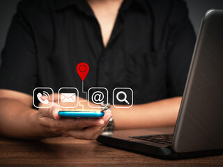 Contact us. Businessman holding a smartphone with a location red pin symbol, search, email, telephone, and address icons while sitting at the table.