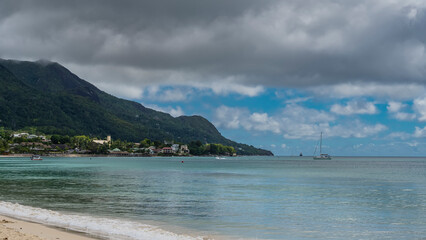 Fototapeta na wymiar The waves of the turquoise ocean are foaming on the sandy beach. Yachts are visible in the distance. A green hill against a background of blue sky and clouds. Seychelles. Mahe. Beau Vallon.