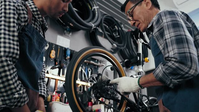 Shopkeeper bicycle senior man training employee to fixed bike in bike shop. Young man assistant and professional repairman assembling bicycle together in cycle store. Maintenance and repair Concept