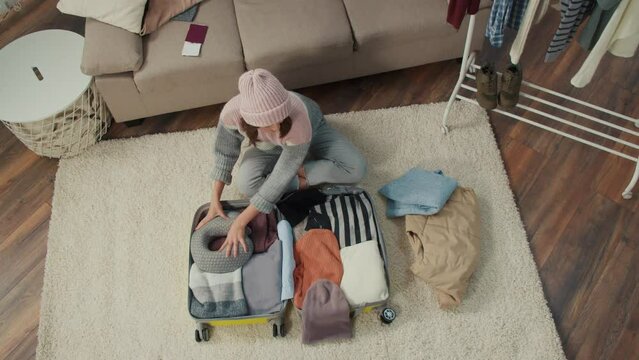 Top View of Young Woman Traveler in a Hat Gathers Clothes for a Trip, Packs Things into a Suitcase at Home. Winter Adventures. Wellness Travel.