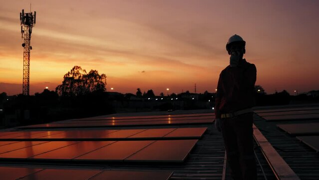Asian technician man using walkie-talkie checking and inform the system solar cell panel on rooftop factory in twilight time, Sustainable energy concept