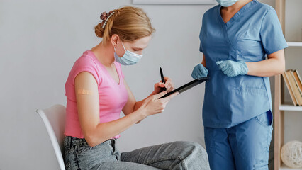Covid-19 vaccination. Medical center policy. Stop pandemic. Young woman in mask with patch after injection on arm sitting on chair signing form and nurse in blue uniform and gloves.