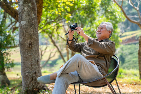 Happy Asian elderly man sitting on outdoor chair and using digital camera taking picture of beautiful nature in public park. Healthy senior man enjoy outdoor lifestyle travel nature on summer vacation
