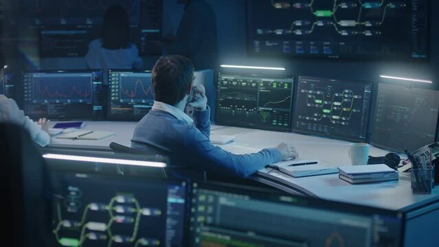 Software engineer works at computer in monitoring room late night. Big data scientists analyze server data. Multiple big screens on the wall with displayed real-time analysis charts. Cyber security.