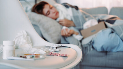 Fever pills. Seasonal flu. Cold remedy. Influenza treatment different medicines sick woman laying on sofa on blur light living room home interior.