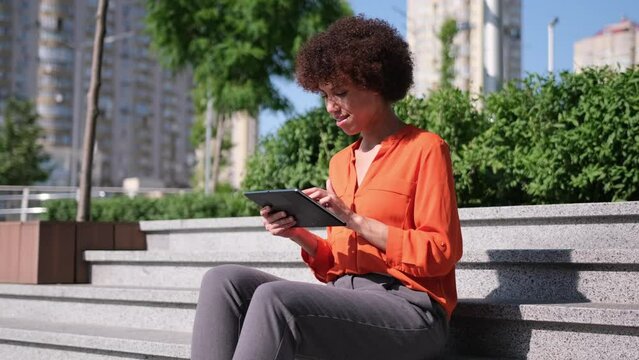 Happy young African American woman using digital tablet pad sufring internet inspired with new ideas sitting outdoors