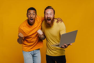 Two excited men expressing winning gesture, celebrating success, holding laptop while standing...