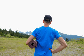 Fototapeta premium Men stand back with yoga mat in mountains landscape. Outdoor yoga in mountains