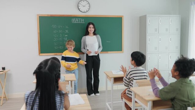 Smart Caucasian cute boy writing answer math formula on blackboard with chalk during lesson at school. Young teacher woman and diverse student clap hands to classmate correct answer in mathematics.